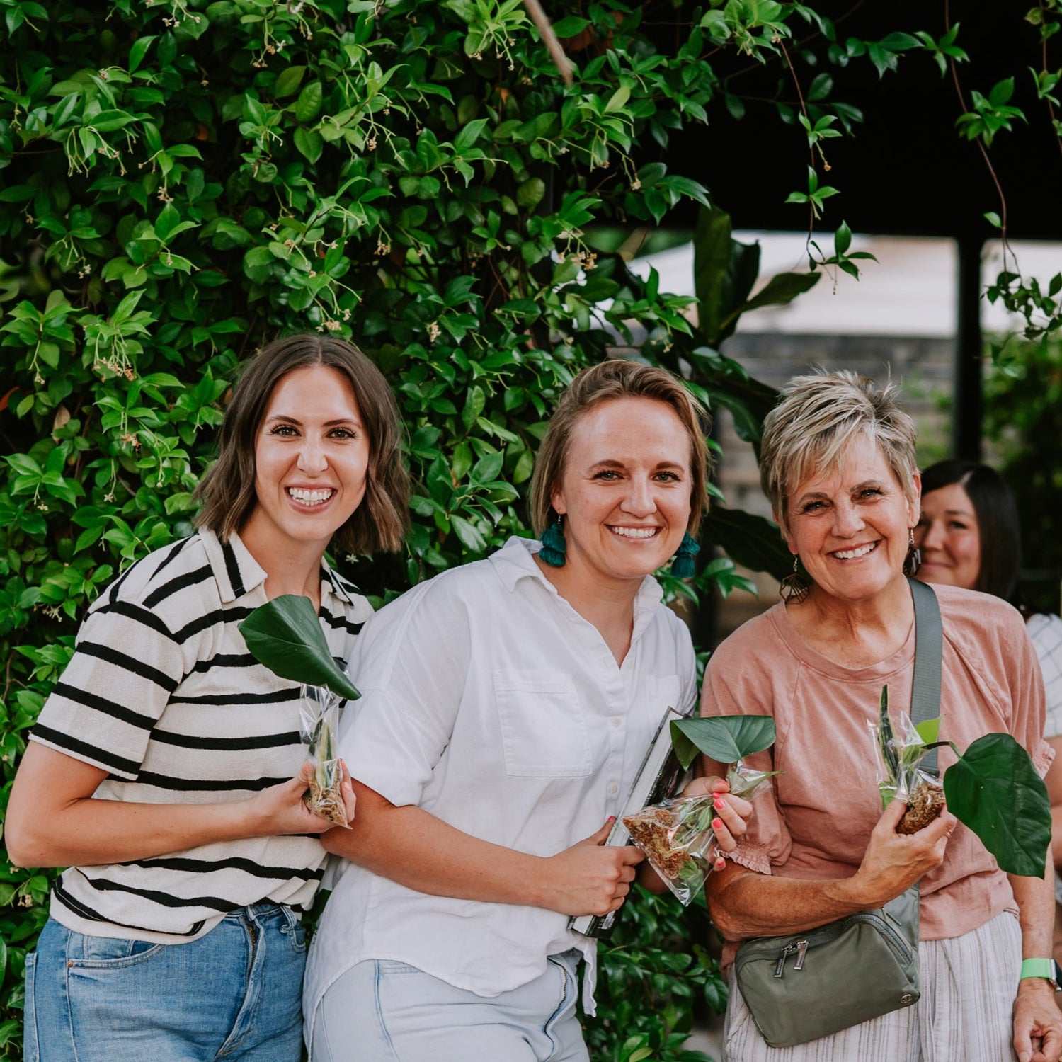 Three women standing together with plants, smiling at the camera against a green leafy background.